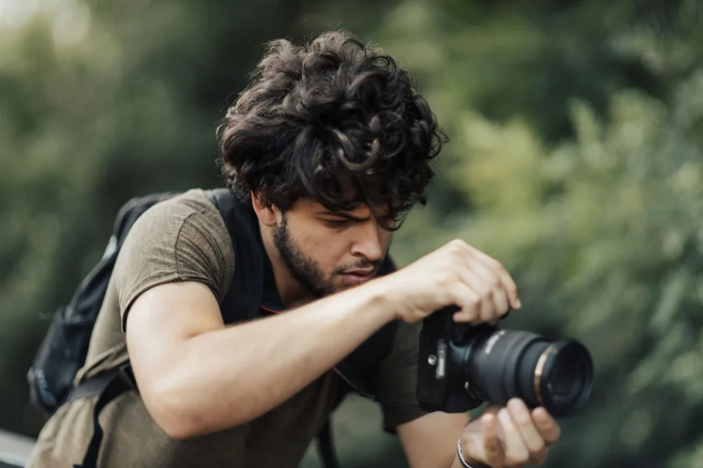 Young photographer in her twenties with curly hair examining photos on her laptop screen, sitting at a modern white desk with editing equipment, warm golden hour light from window, eye-level medium shot, lifestyle photography, shallow depth of field