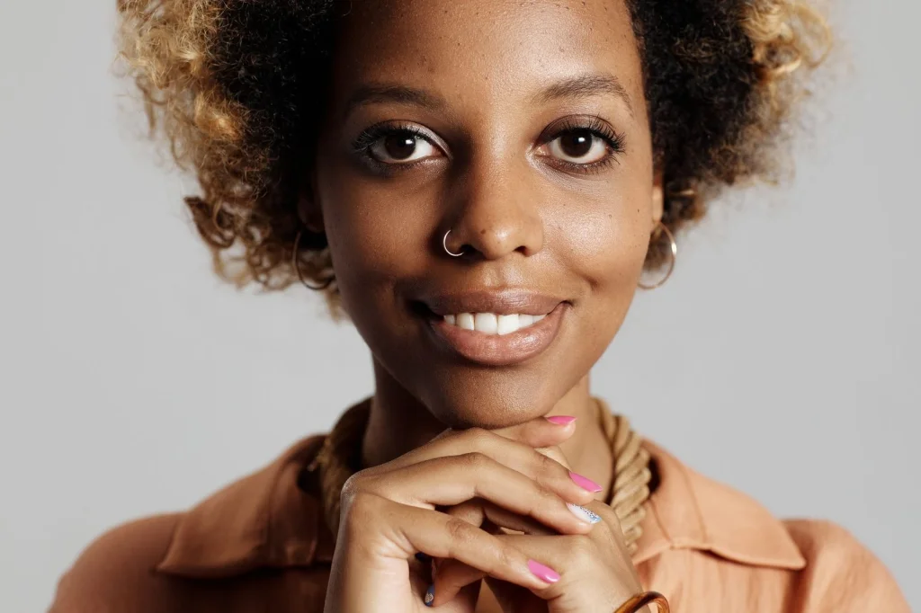 Young Black woman with natural 4B hair in a cream colored top, looking frustrated while using phone, sitting on a gray couch in modern living room, soft window light from left side, medium shot at eye level, lifestyle photography, natural depth of field