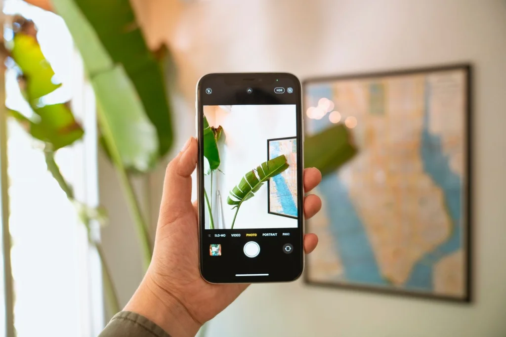 Close-up of hands holding a smartphone showing a photo editing app interface, sitting at a white desk with coffee cup and notebook, natural daylight from window, overhead angle, lifestyle photography