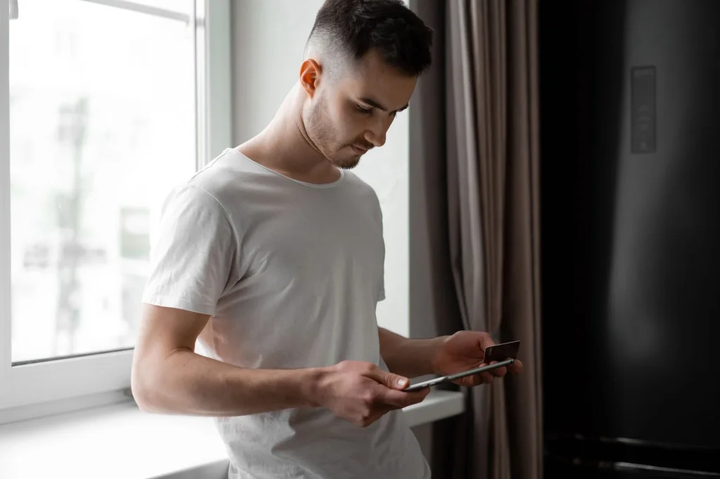 Young man in casual clothing holding a tablet showing face swap app results, modern living room background, warm ambient lighting, over-shoulder view, lifestyle photography, natural depth of field
