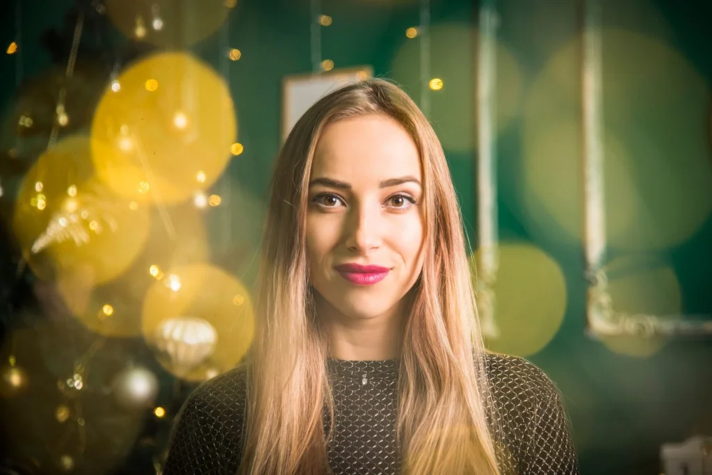 Professional woman in her late 20s with shoulder-length blonde hair, subtle smile, wearing a navy blue blazer over white blouse, sitting at a modern office desk with plants in background, soft natural light from large window, eye-level medium shot, editorial portrait, shallow depth of field