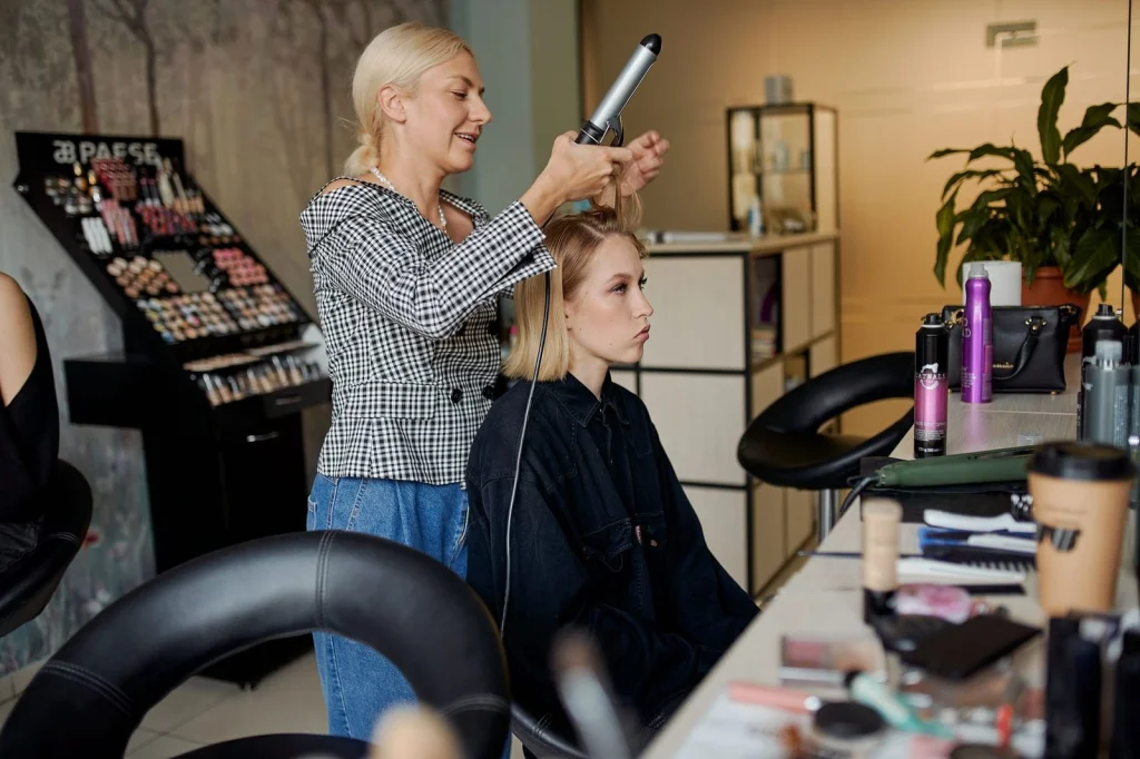 Professional hairstylist in her 30s with blonde hair in a sleek bob, holding styling tools, standing in a modern salon with mirrors and good lighting, natural daylight mixed with salon lighting, medium shot showing upper body, editorial photography, professional atmosphere