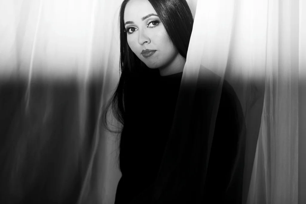Woman in her 30s with long dark hair, white blouse, slight smile, holding a tablet showing a product photo with transparent background, sitting at a modern white desk in a bright office, soft window light from the left, eye-level medium shot, lifestyle photography, shallow depth of field