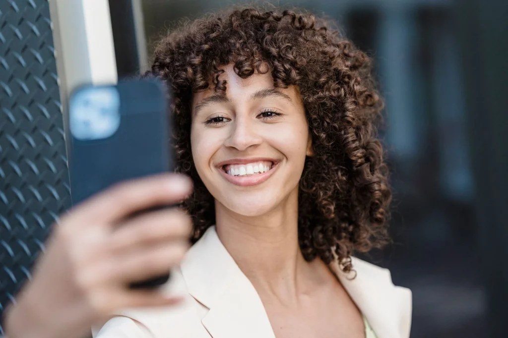 Young woman with voluminous curly brown hair, natural smile, wearing a white blouse, standing against a transparent checkered background pattern, studio lighting from above, three-quarter view portrait, commercial photography, crisp focus