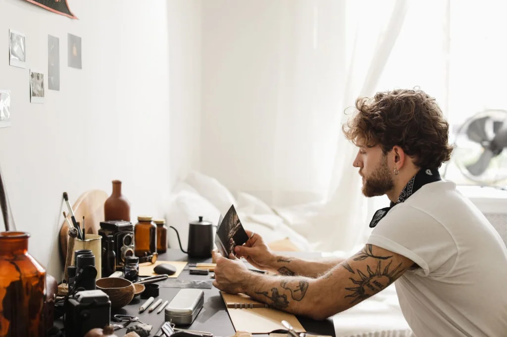 Professional photographer in his 30s with beard sitting at a modern workspace, editing product photos on a large monitor, multiple camera lenses on the desk, warm LED desk lighting, over-shoulder medium shot, editorial workplace photography