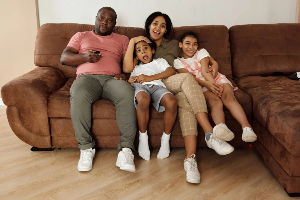 Family of four sitting together on a beige couch, parents in their 40s and two children aged 8-12, all wearing casual clothing and smiling at the camera, bright living room with white walls, soft afternoon lighting from large windows, medium wide shot at eye level, family portrait photography, warm and inviting