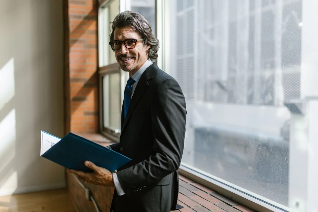 Businessman in his 40s wearing a dark suit and blue tie, confident expression, sitting at a modern office desk with computer monitors, soft overhead lighting, medium shot, corporate portrait