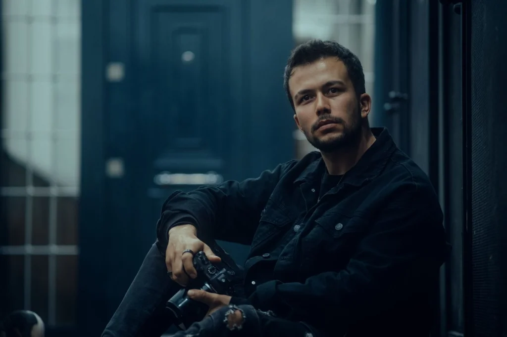 Professional photographer in his 30s with stubble, focused expression, sitting at a modern desk with laptop and multiple photo editing tools open on screen, soft office lighting from window, eye-level medium shot, editorial portrait, shallow depth of field
