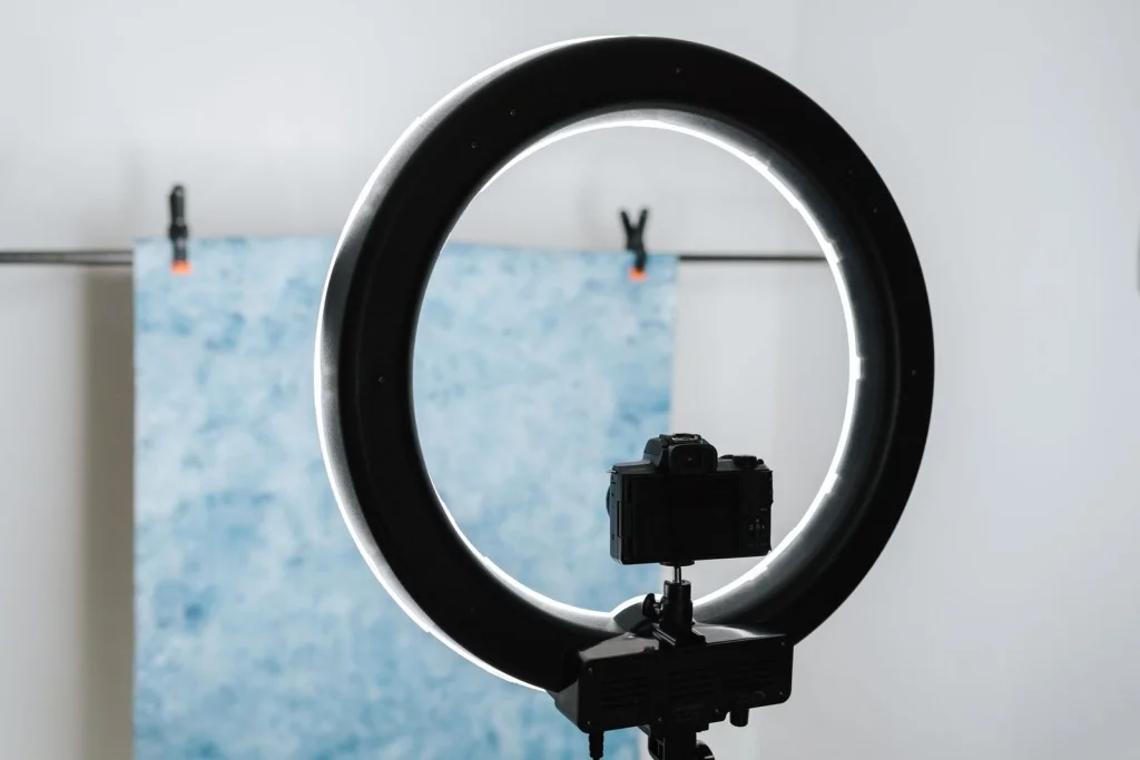 Professional camera setup with ring light and white backdrop in photography studio, Canon camera on tripod, model in background, warm studio lighting, wide establishing shot, behind-the-scenes photography