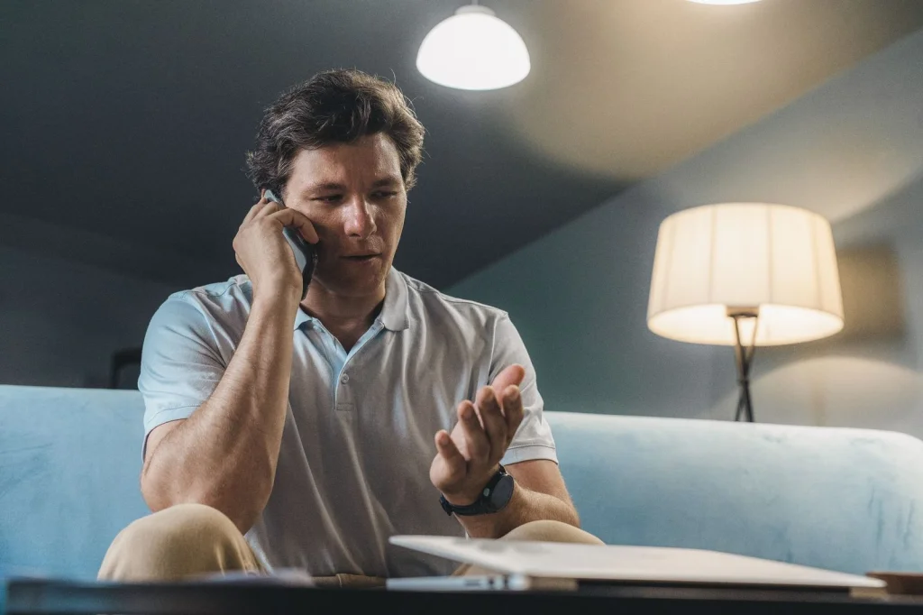 Man in his 30s with short brown hair comparing photo editing results on a dual monitor setup, looking focused while scrolling through before and after images, sitting at a modern white desk with a mechanical keyboard, warm desk lamp lighting from the right, overhead three-quarter view, tech workspace photography