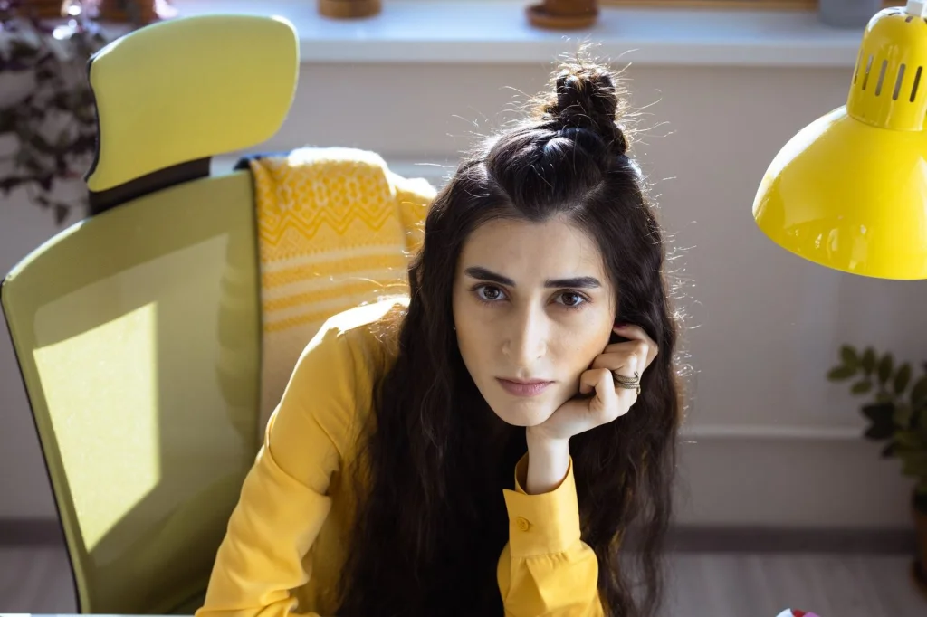Woman in her 20s with curly hair sitting at a desk, looking frustrated while working on a computer with Photoshop interface visible on screen, home office setting with plants in background, natural window light from the right, medium shot from side angle, lifestyle photography, shallow depth of field