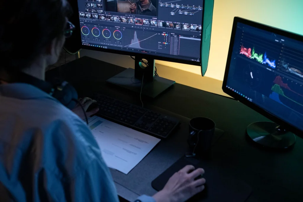 Designer working on product photography editing, removing backgrounds from multiple shoe images on dual monitors, modern studio setup with professional lighting equipment, side angle shot, commercial photography workspace