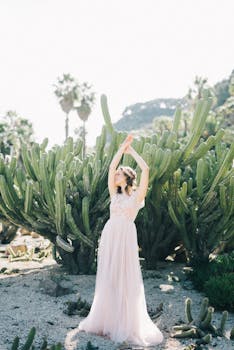 A woman in a flowing white dress poses gracefully among cacti in a sunny garden.